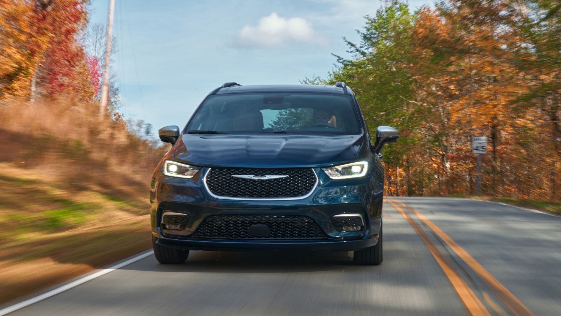 Dynamic front-end shot of a dark blue 2025 Chrysler Pacifica Plug-in Hybrid Pinnacle driving on a country road with trees in the background.