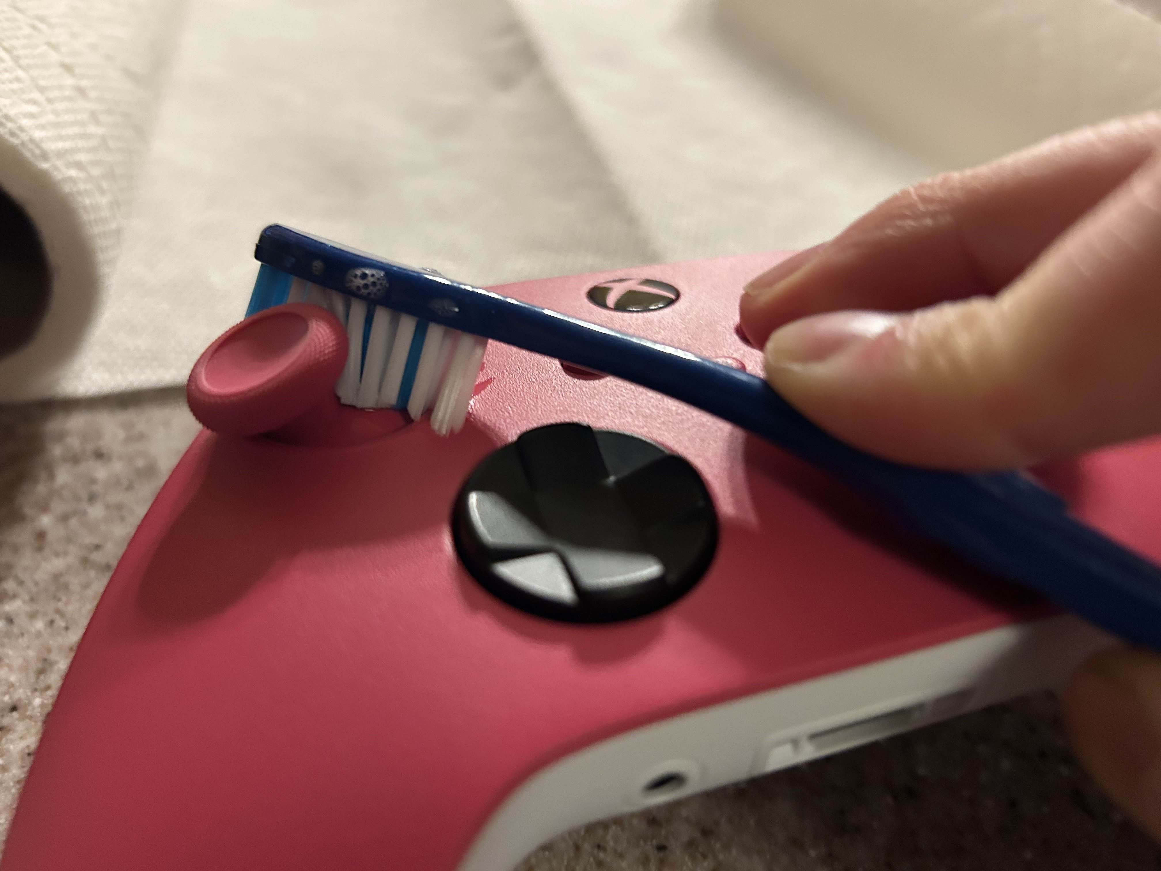 Cleaning the Side of a Joystick of a Pink Xbox One Controller with a Blue Toothbrush