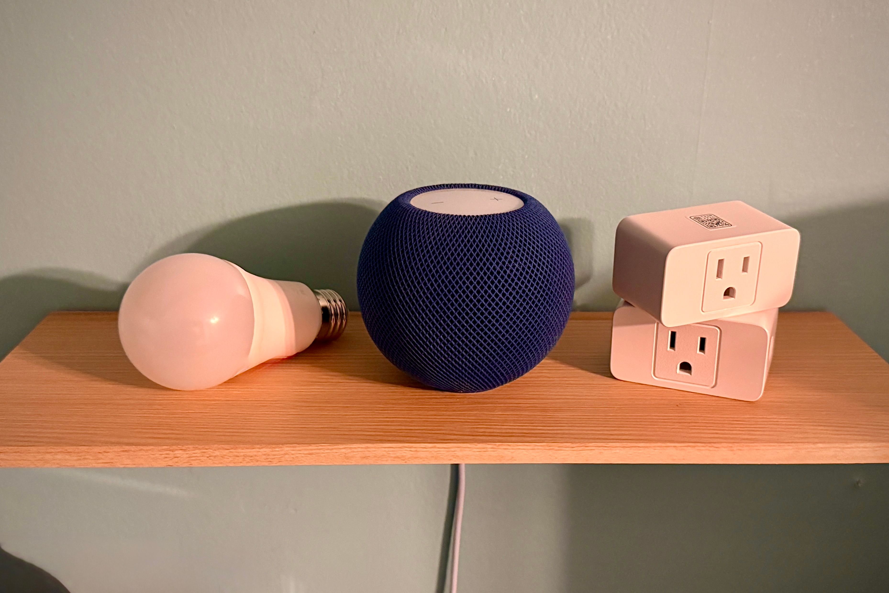 Bleu HomePod Mini ao lado de dois plugues inteligentes e uma lâmpada inteligente em uma prateleira.