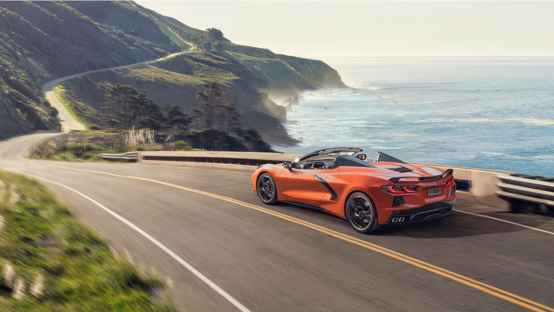 Dynamic rear 3/4 shot of an orange 2020 Chevrolet Corvette Stingray Convertible driving on a country road running beside the ocean.