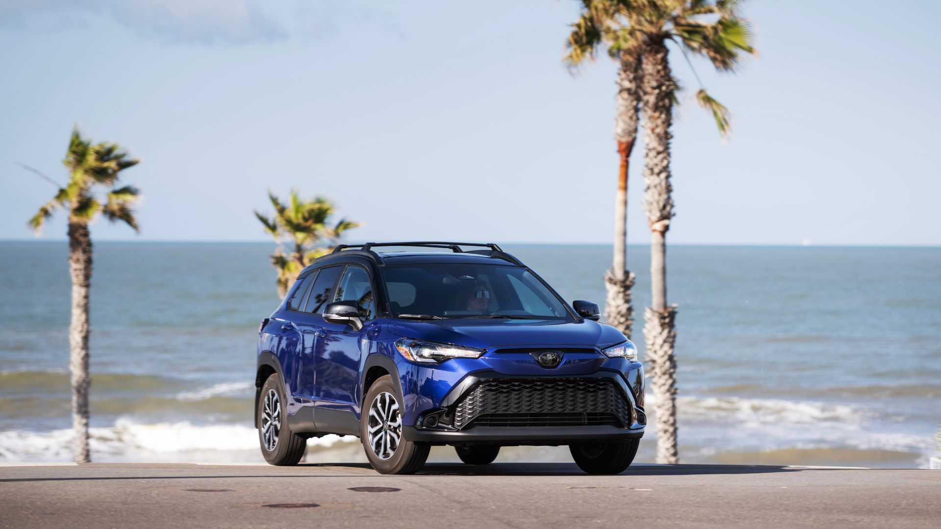 Static Front 3/4 Shot of a Blue 2024 Toyota Corolla Cross Hybrid SE with palm trees and ocean in background.