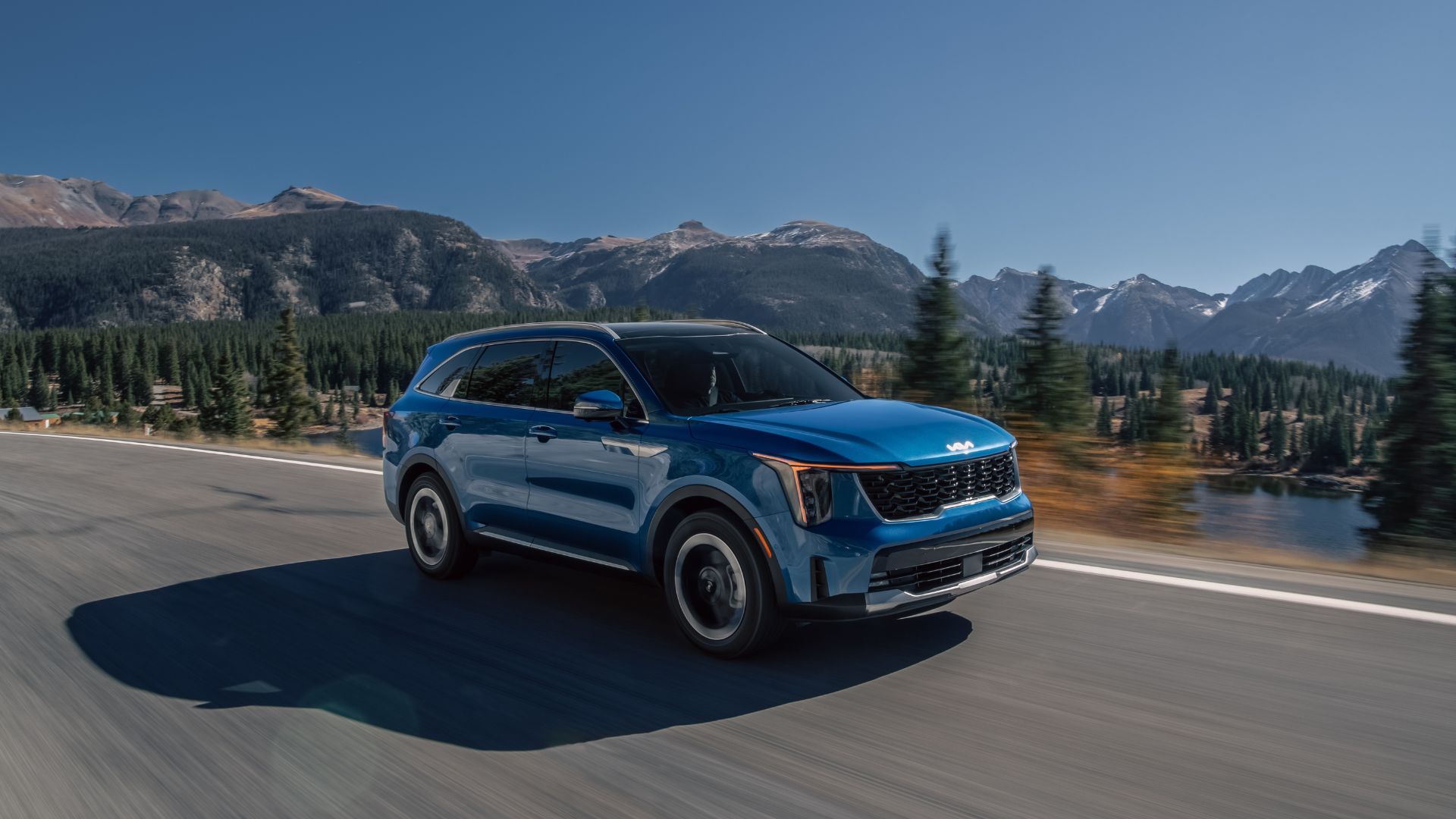 Dynamic front 3/4 shot of a blue 2025 Kia Sorento HEV driving on a country road with snow-capped mountains in the background.