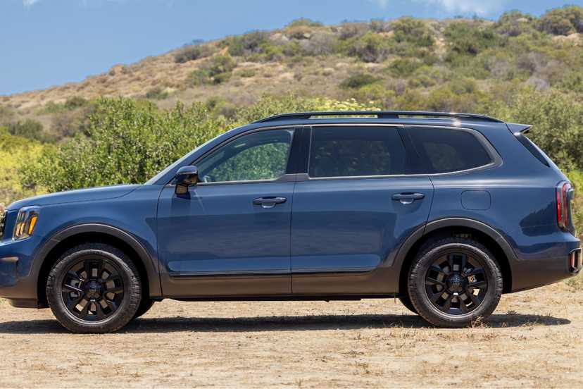 Static side profile shot of a blue 2025 Kia Telluride parked on sand with a grassy hill in the background.