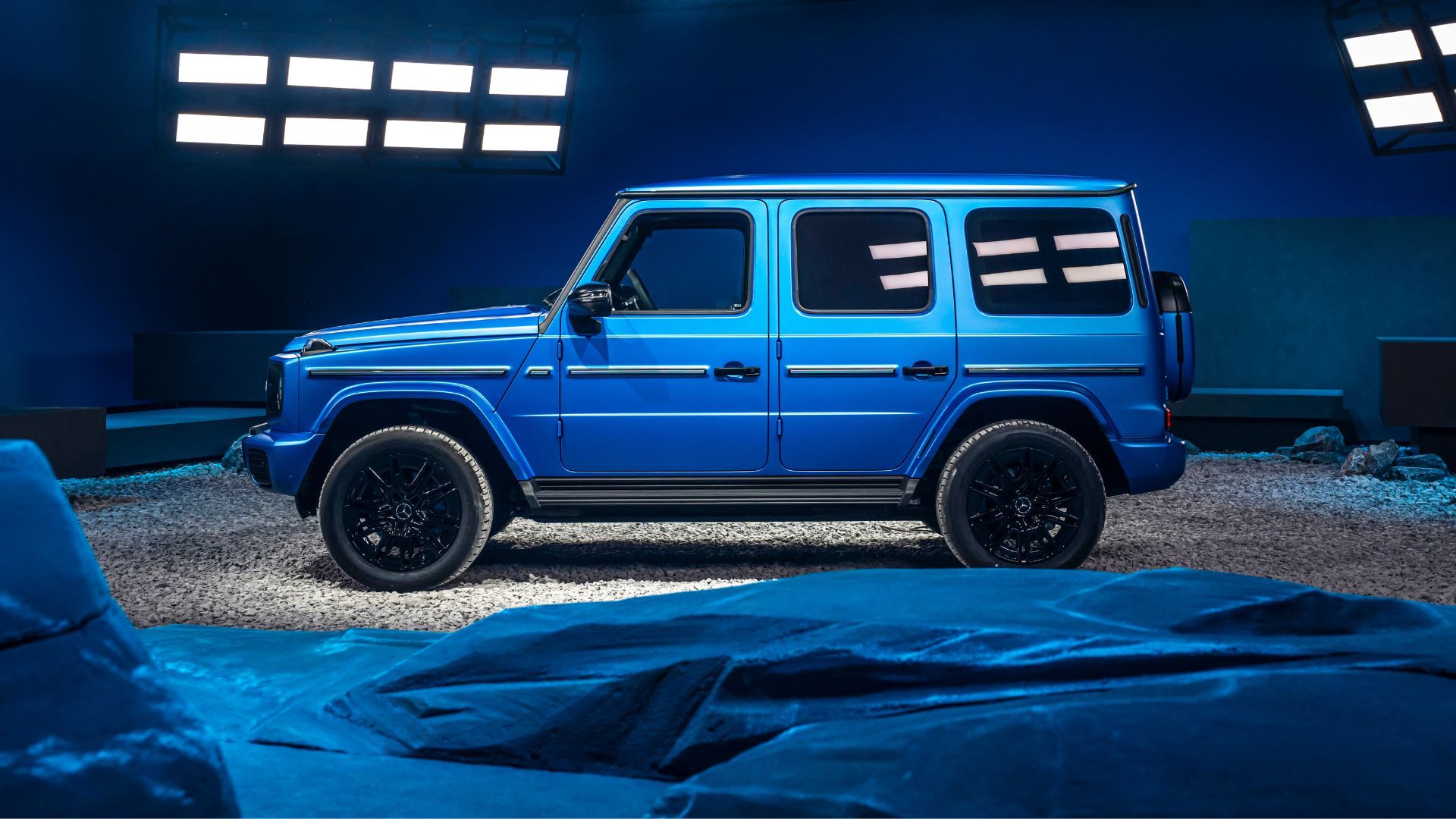 Static side profile shot of a blue Mercedes-Benz G580 EQ in a studio setting.