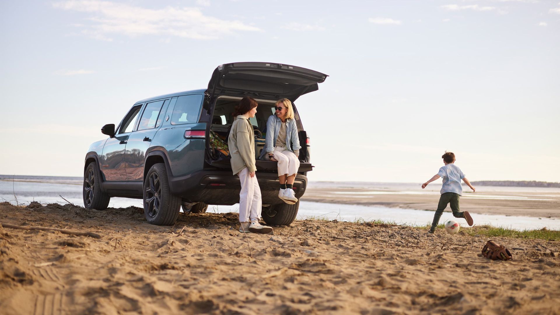 Static rear 3/4 shot of a Rivian R1S parked oin a beach with two people stood at the rear of its open trunk.