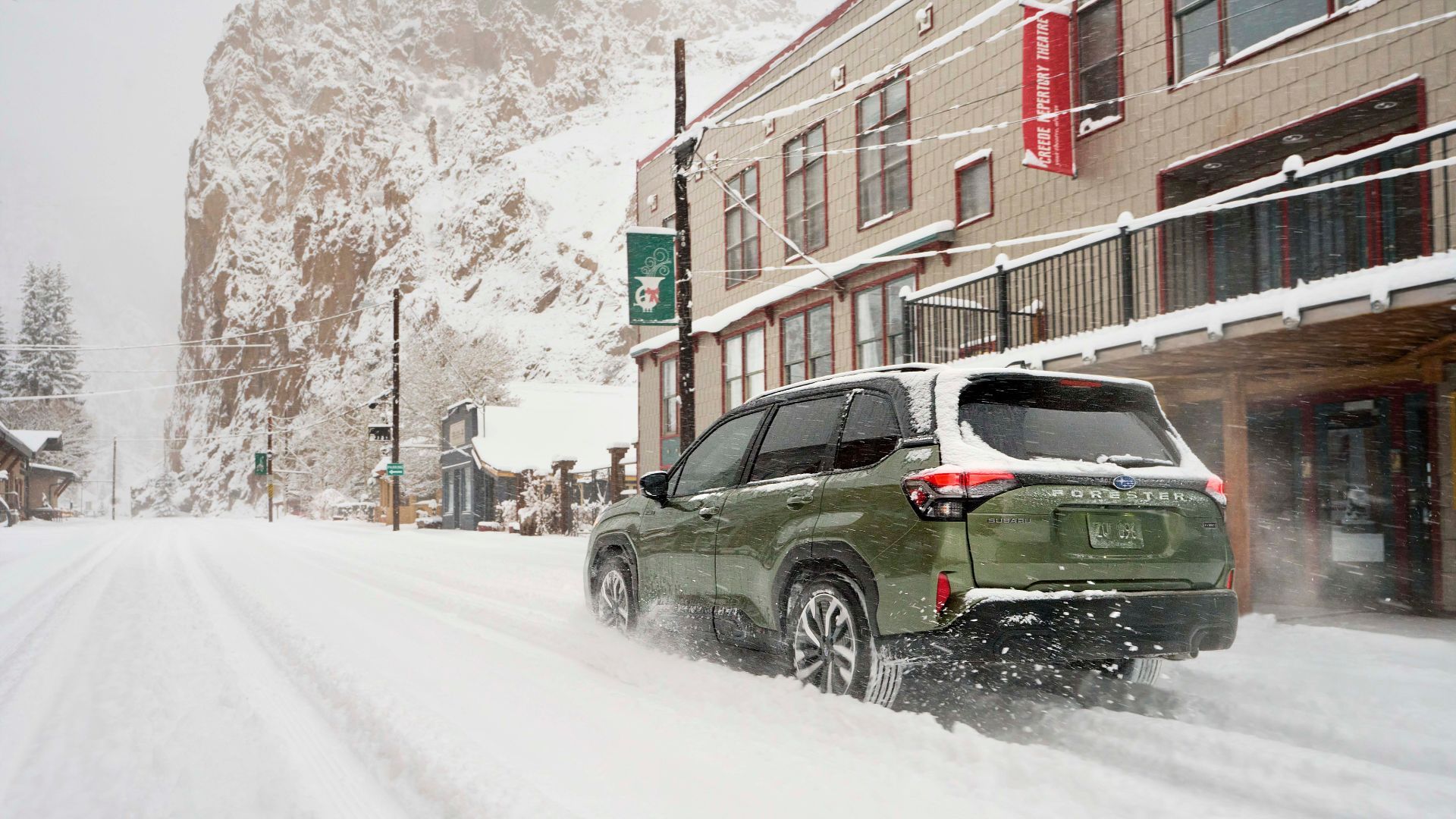 Dynamic rear 3/4 shot of a green 2025 Subaru Forester Hybrid driving in snow in a city.