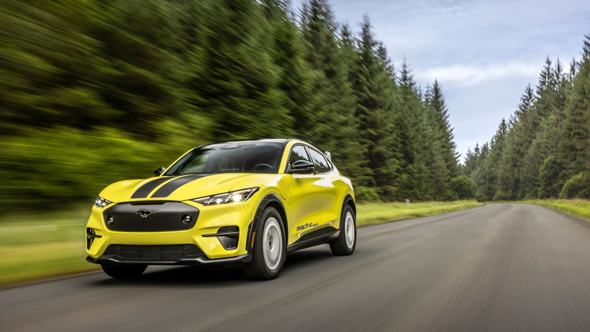 Dynamic front 3/4 shot of a yellow Ford Mustang Mach-E Rally driving on a gravel track through a forest.