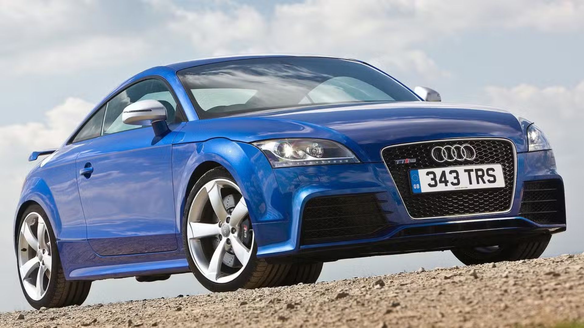 Static front 3/4 shot of a blue Audi TT RS parked on gravel.