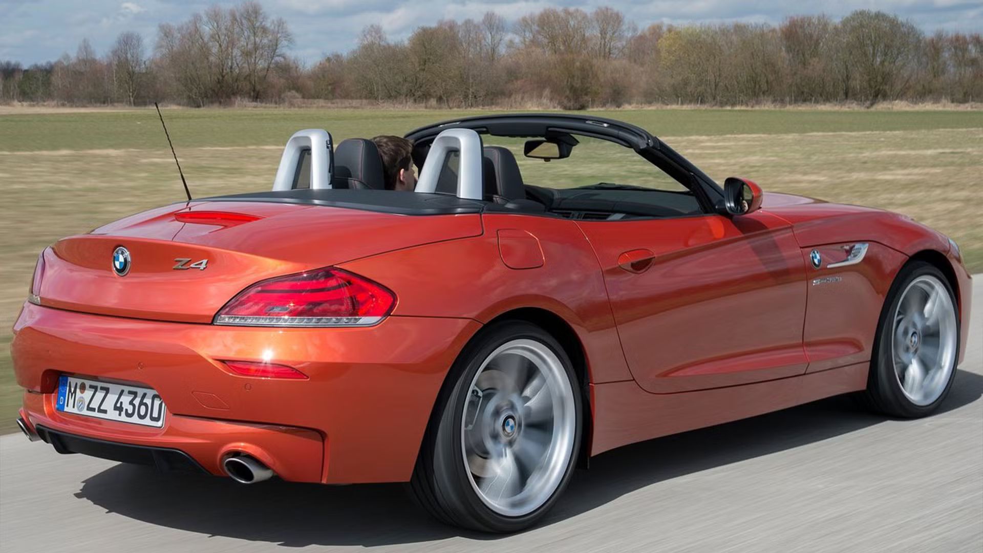Dynamic rear 3/4 shot of an orange BMW Z4 driving with its roof down on a country road.