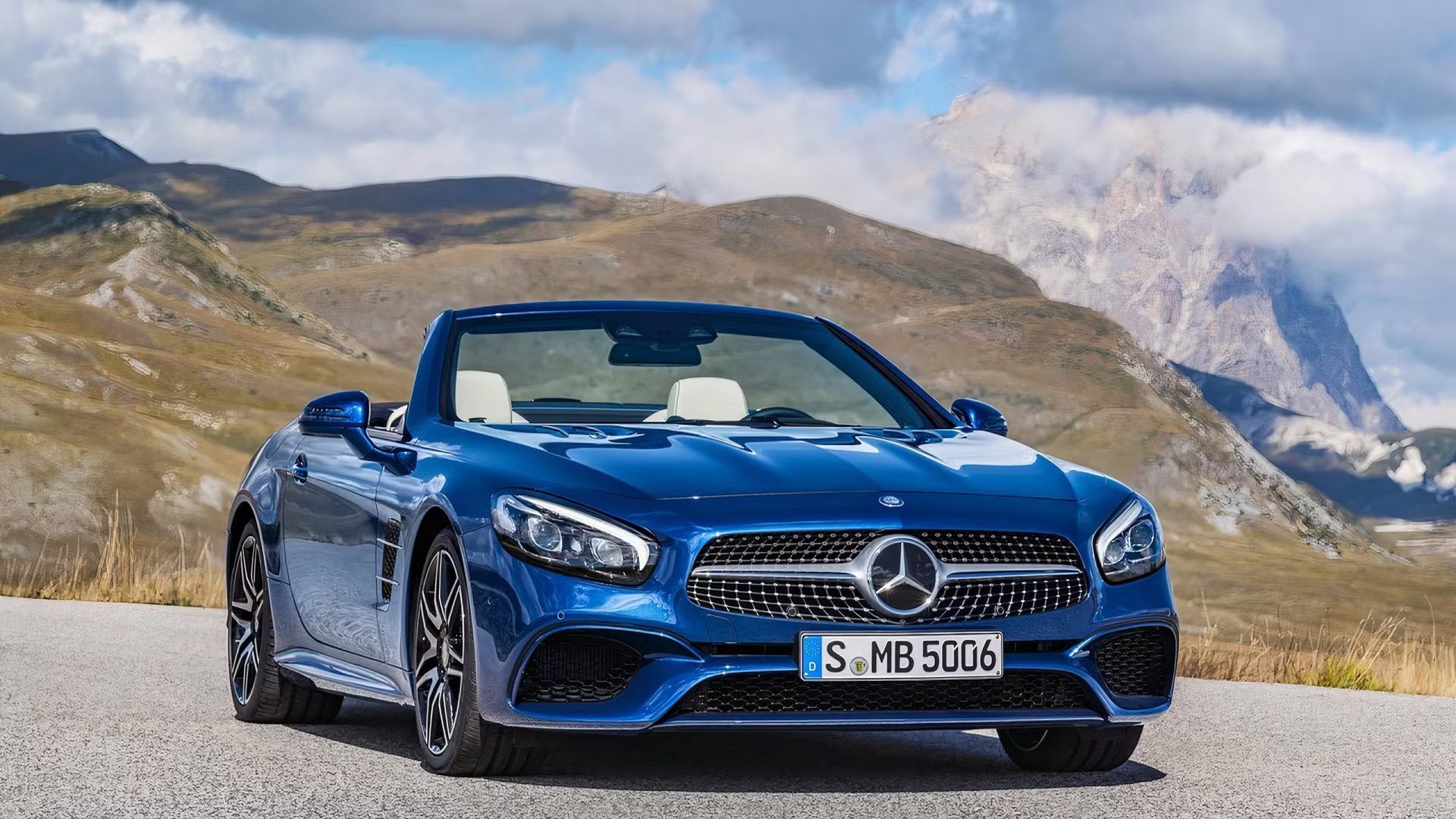 Static front 3/4 shot of a blue Mercedes-Benz SL-Class parked on tarmac with its roof down and mountains in the background.