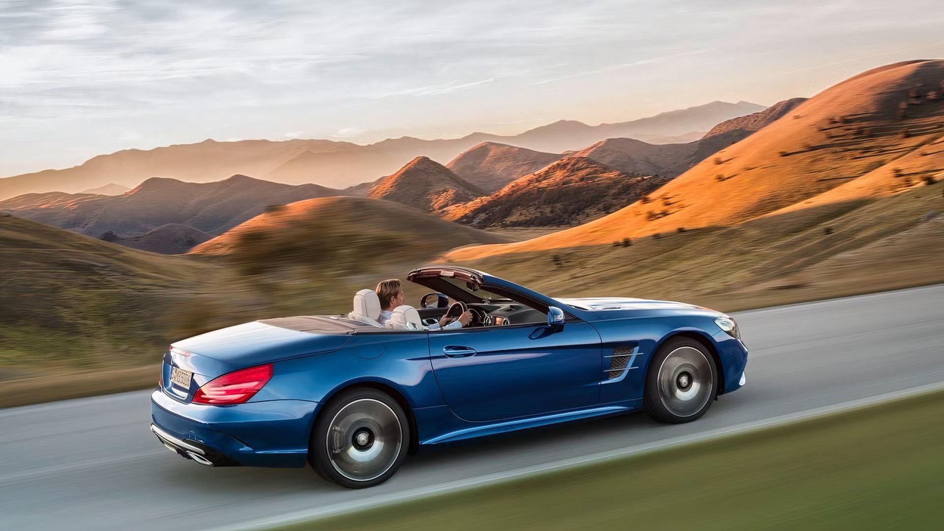 Dynamic rear 4/3 shot of a blue Mercedes-Benz SL-Class with its roof down driving on a country road with mountains in the background.