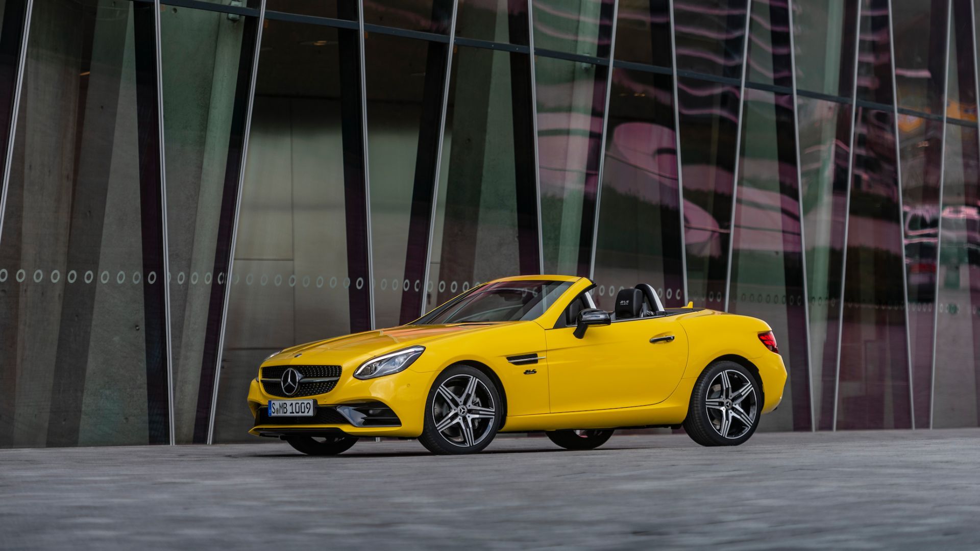 Static front 3/4 shot of a yellow 2020 Mercedes-Benz SLC with its roof folded down parked outside of a modern building.