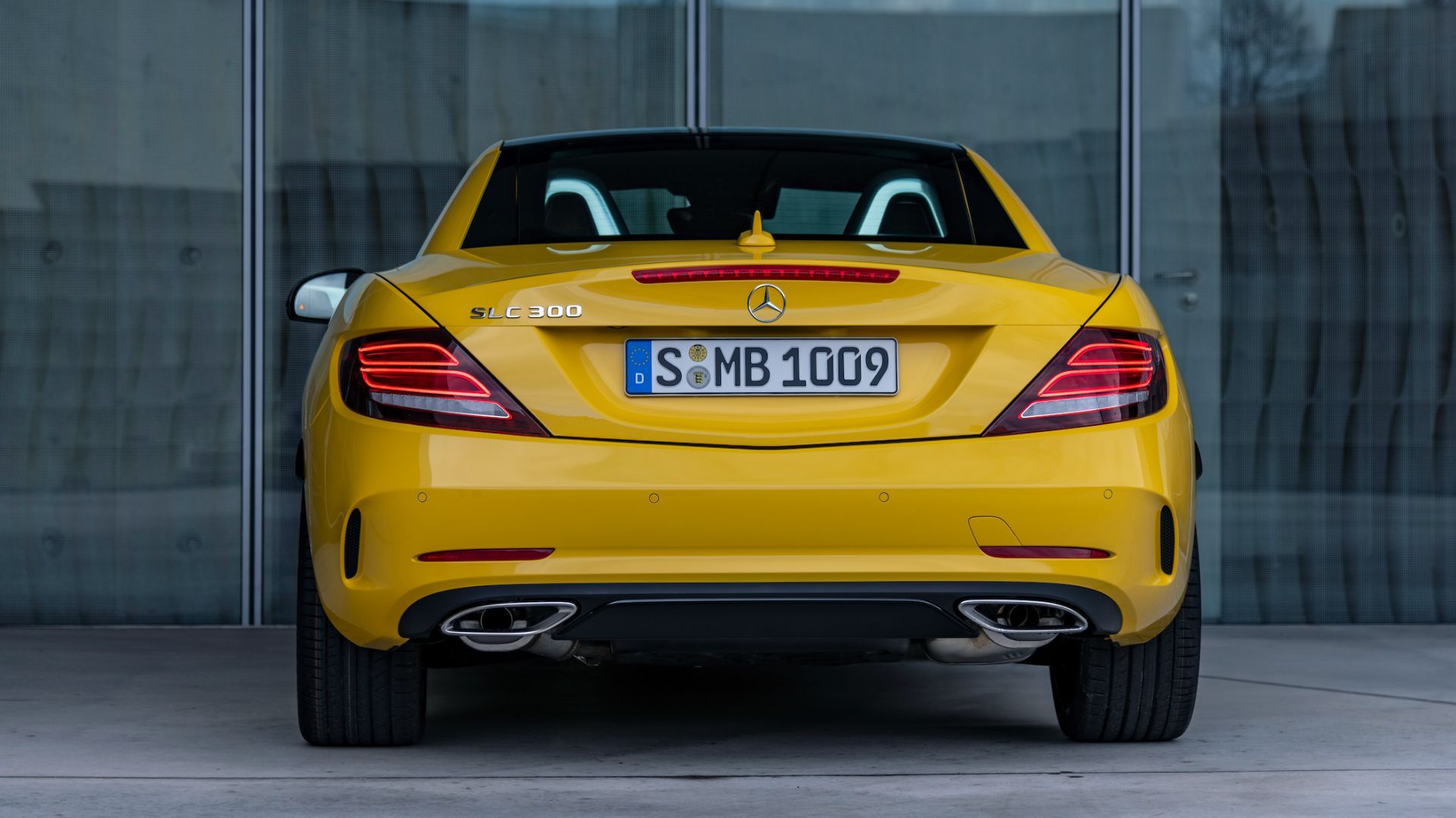 Static rear end shot of a yellow 2020 Mercedes-Benz SLC with its roof down parked outside of a modern building.