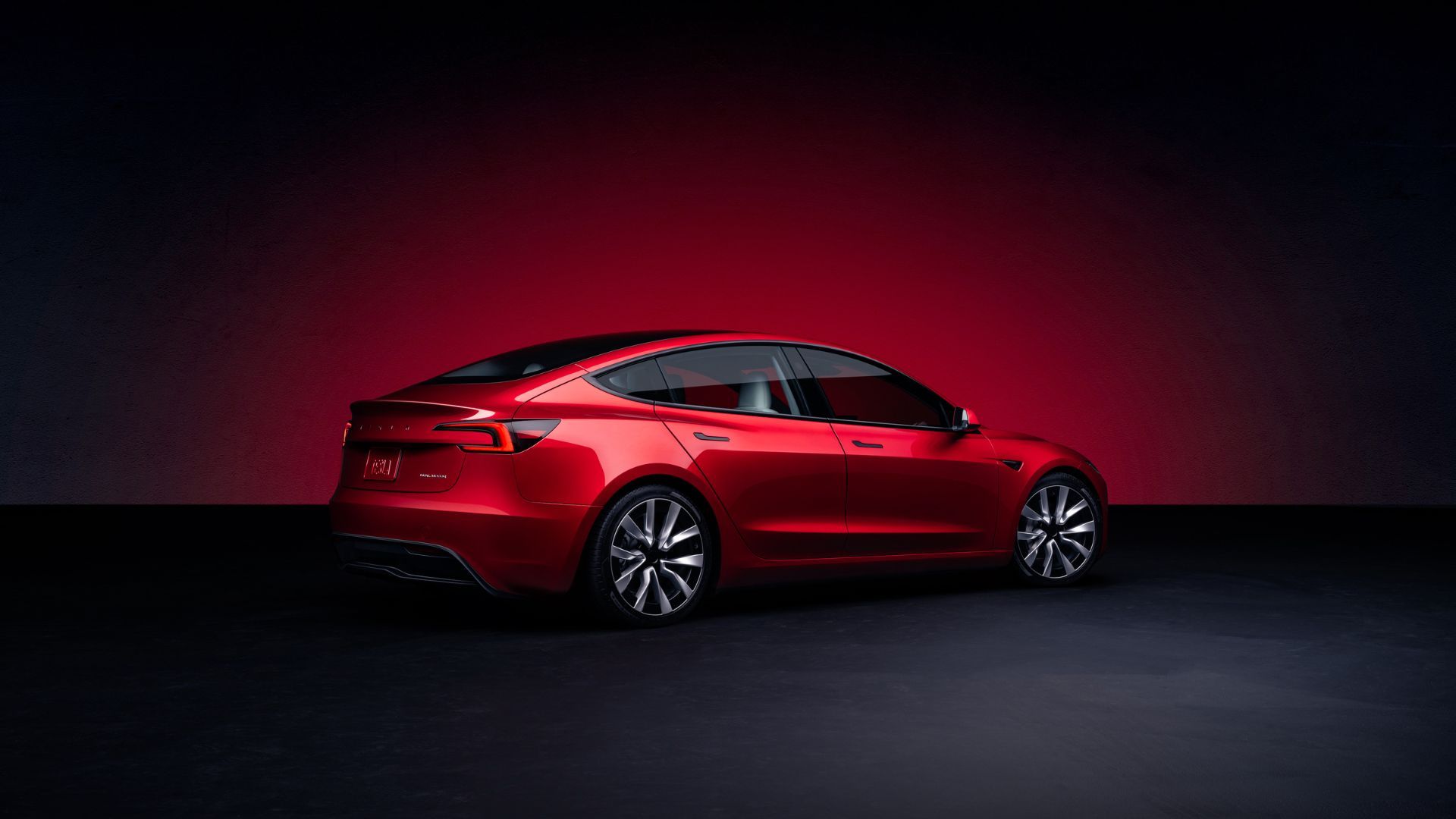 Static rear 3/4 shot of a red Tesla Model 3 in a studio with a red and black backdrop.