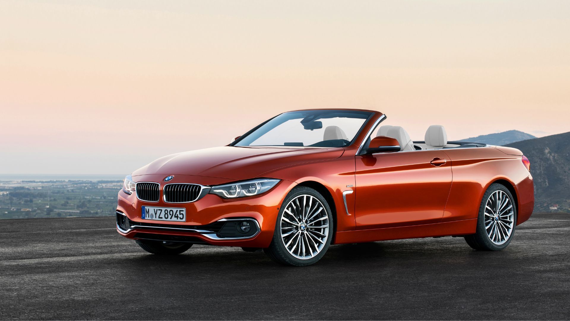 Static front 3/4 shot of an orange 2017 BMW 430i Convertible parked on tarmac with a cityscape in the background.