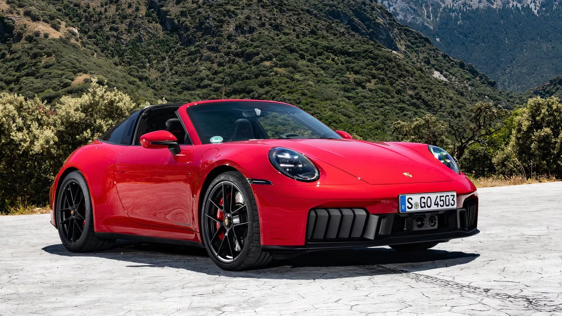 Static front 3/4 shot of a red Porsche 911 Targa parked on tarmac with a mountain in the background.