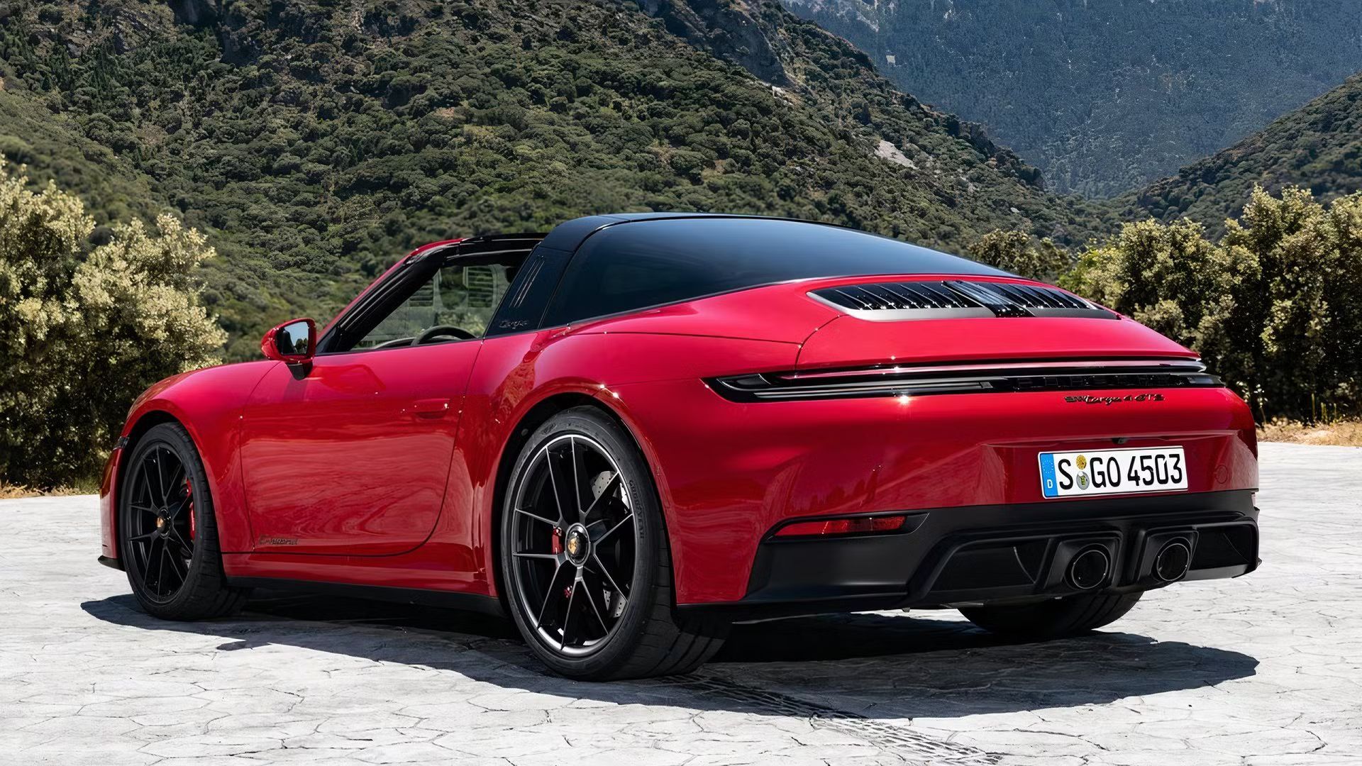 Static rear 3/4 shot of a red Porsche 911 Targa parked on tarmac with a mountain in the background.