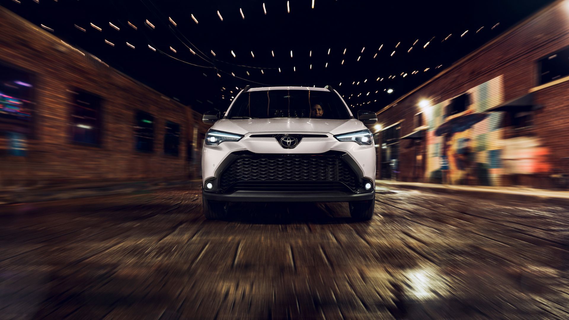 A white 2025 Toyota Corolla Cross Hybrid Dynamic Front-Shot driving on a cobble street in a city at night.