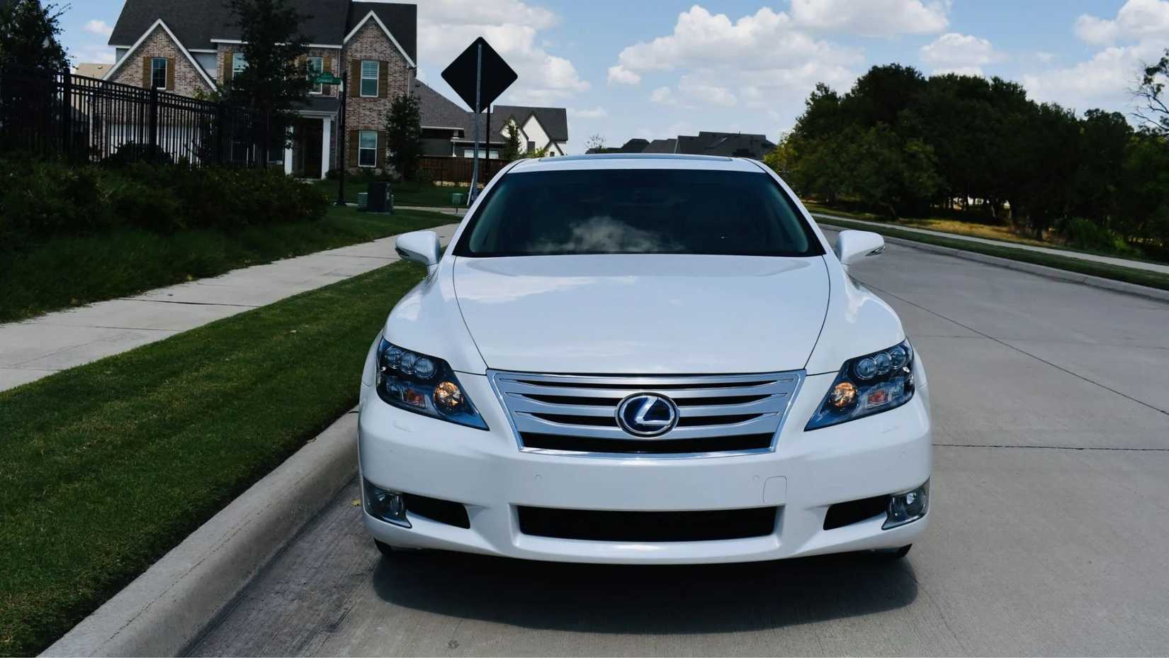 Static front-end shot of a white 2011 Lexus LS 600h L parked next to a sidewalk with houses in the background.