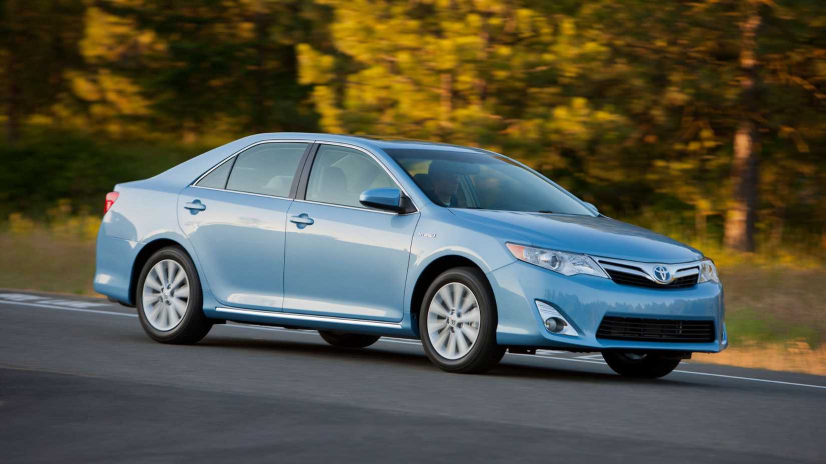 Dynamic front 3/4 shot of a blue 2012 Toyota Camry Hybrid driving on a road with trees in the background.