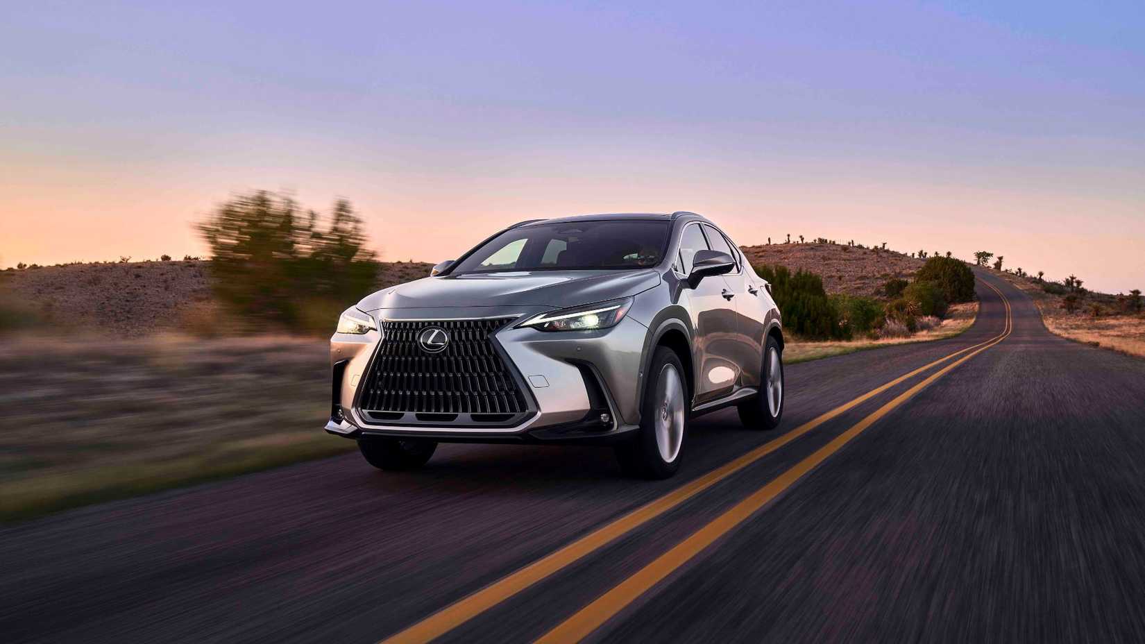 Dynamic front 3/4 shot of a gray 2025 Lexus NX 350h driving on a country road at sunset.