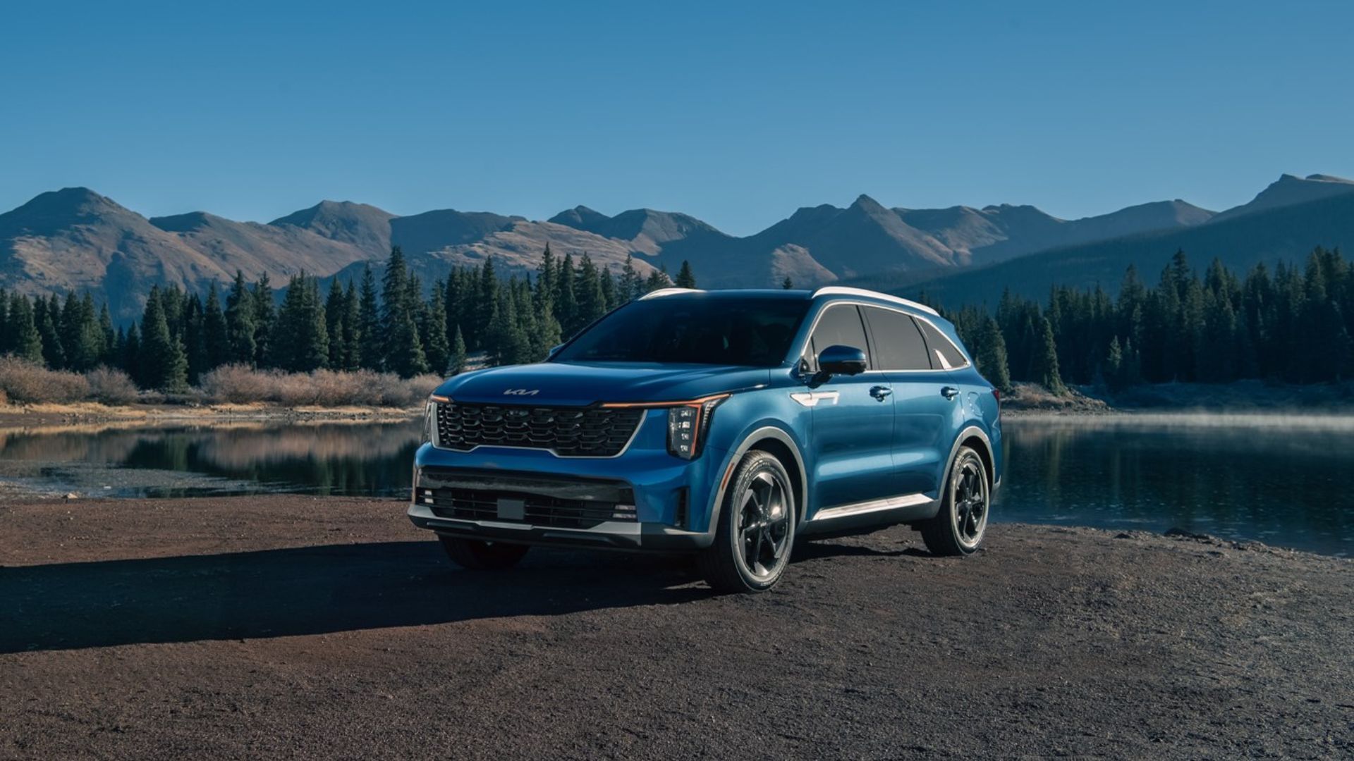Static front 3/4 shot of a blue 2025 Kia Sorento Hybrid parked next to a lake with mountains in the background.
