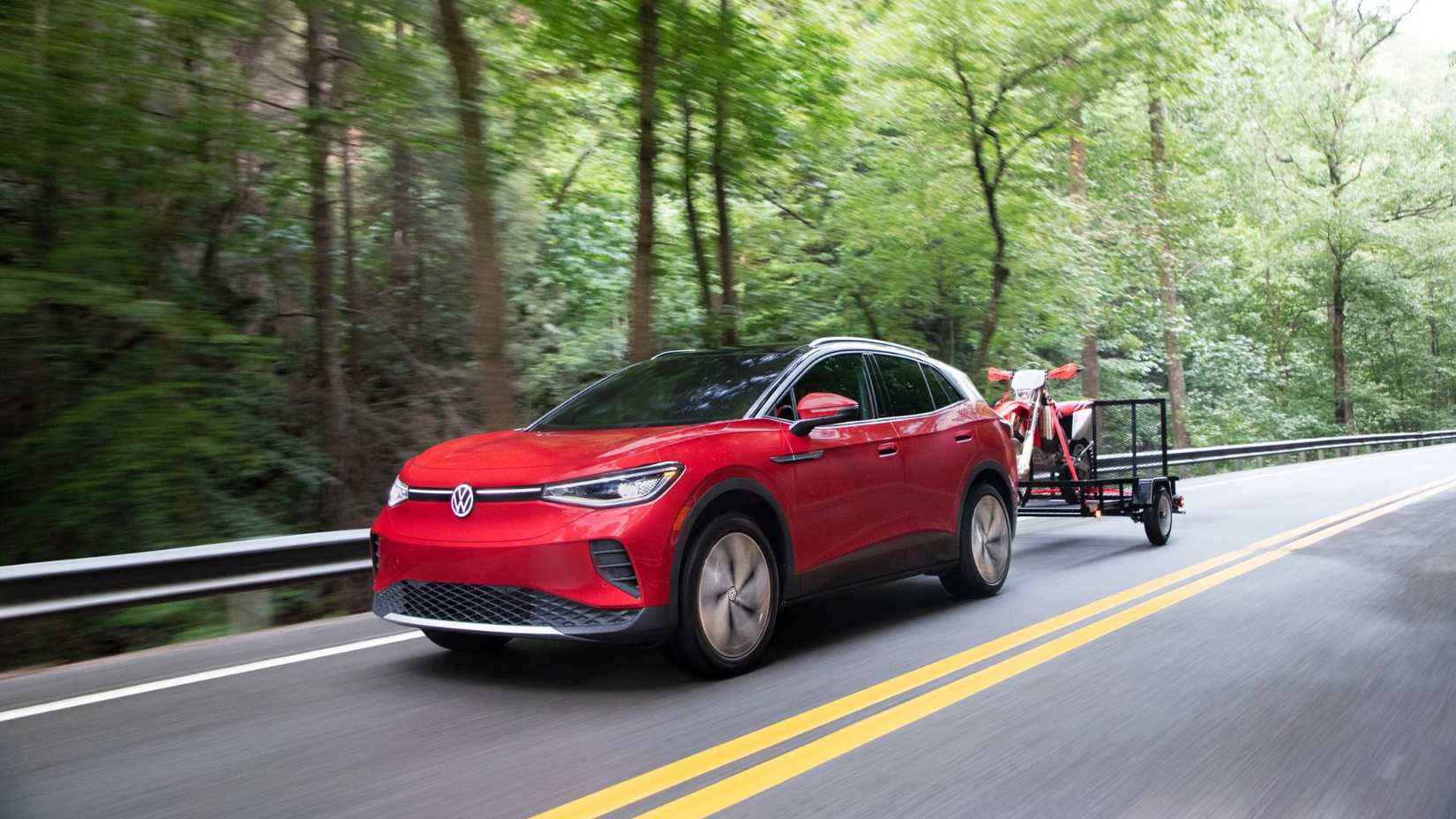 Dynamic front 3/4 shot of a red 2021 Volkwagen ID.4 towing a trailer carrying a dirt bike on a country road lined by trees.