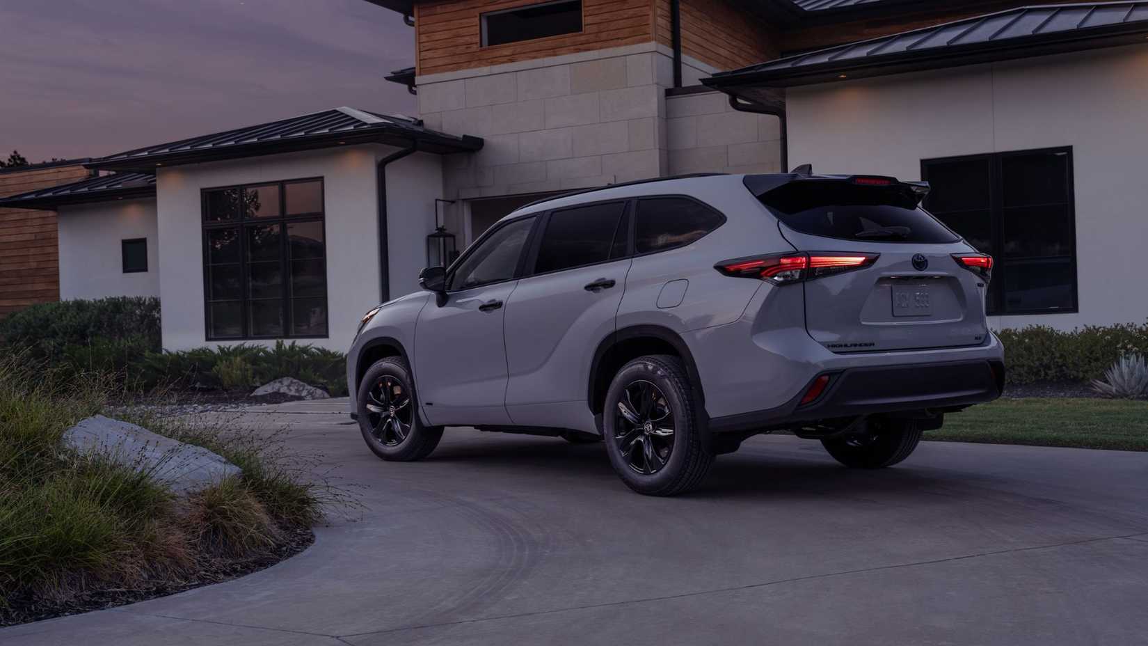 Static rear 3/4 shot of a gray 2025 Toyota Highlander Hybrid parked on the driveway of a house.
