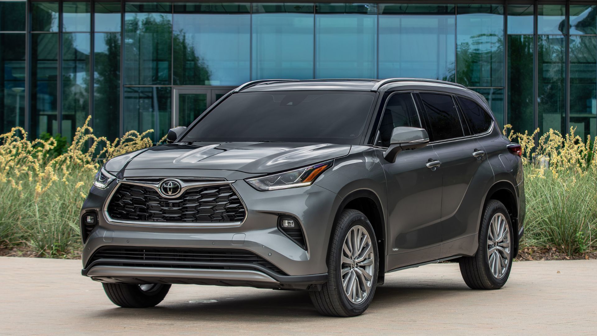 2Static front 3/4 shot of a gray 2025 Toyota Highlander Hybrid parked outside of a glass-fronted building.