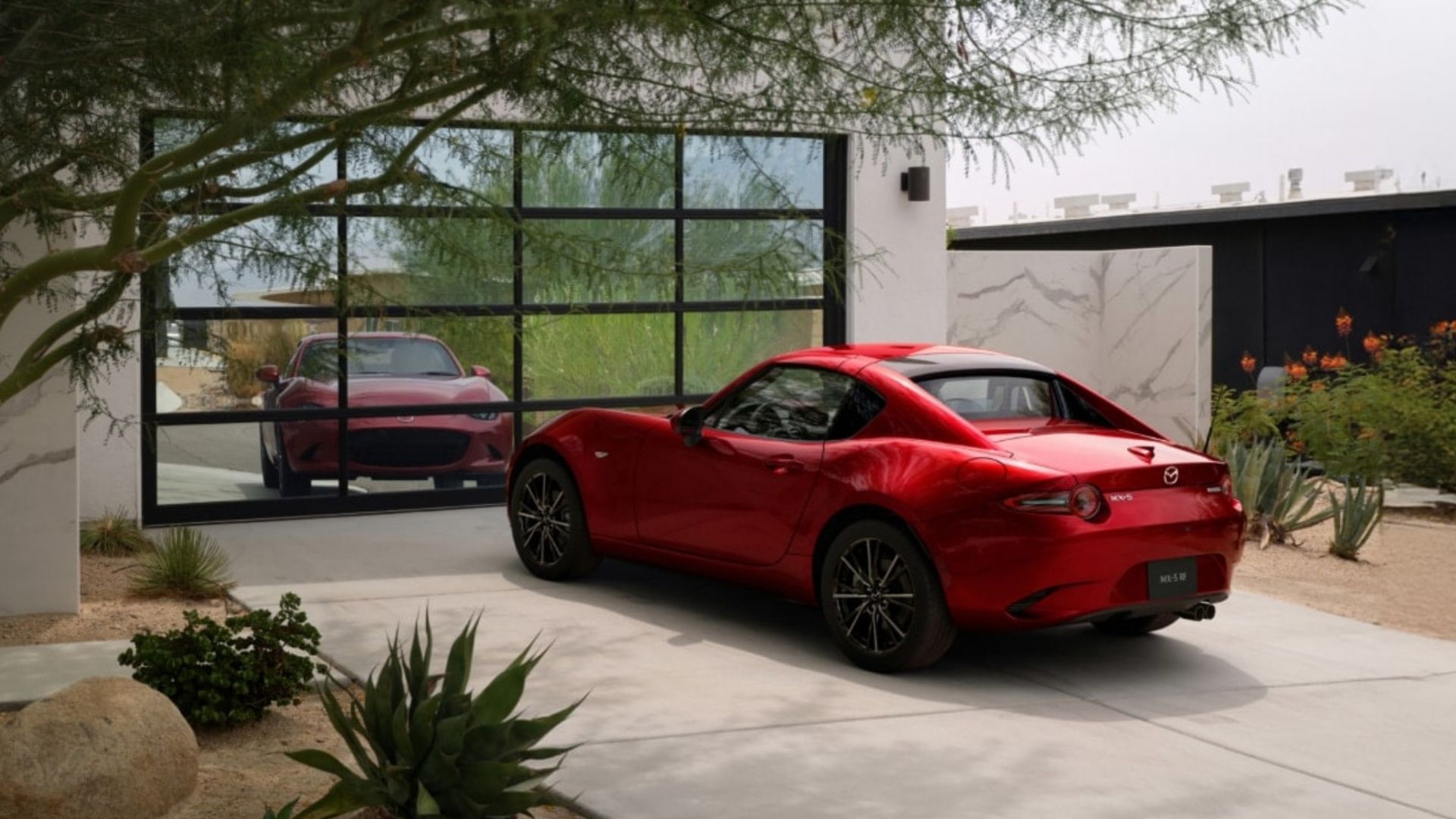 Static rear 3/4 shot of a red 2025 Mazda MX-5 Miata RF parked on the driveway of a house with its roof up.
