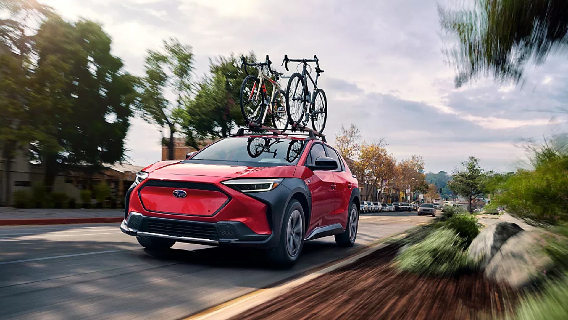 Dynamic front 3/4 shot of a red 2025 Subaru Solterra driving on a country road with bikes on its roof.