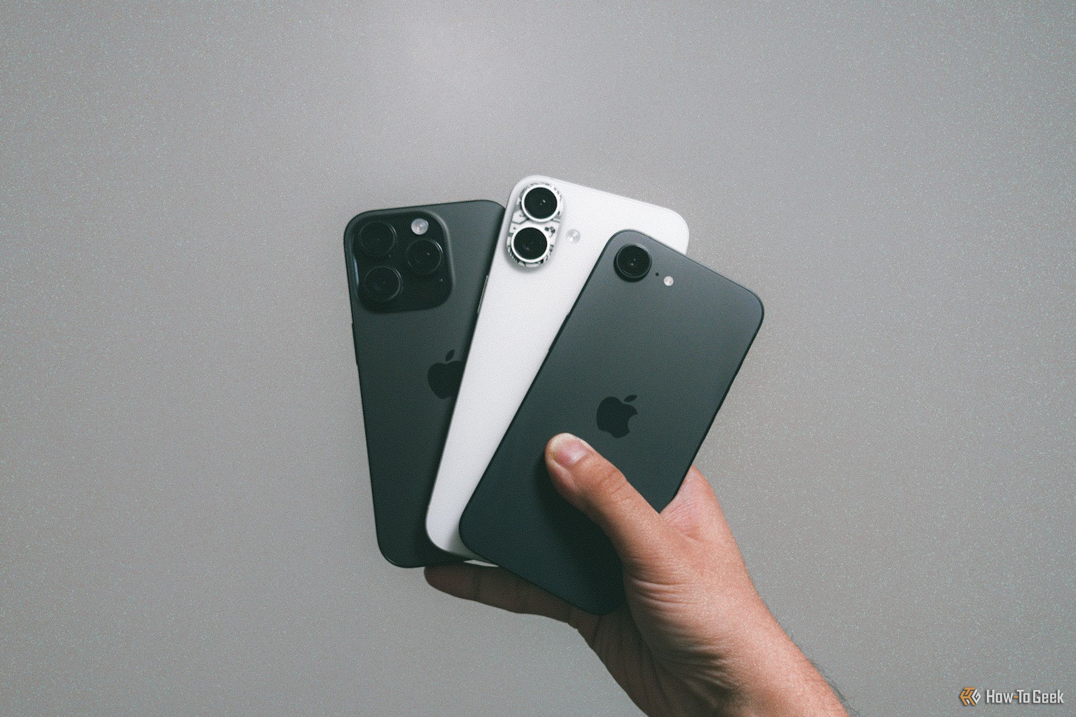 iPhone 16e with iPhone 16 Plus and iPhone 16 Pro in hand against grey wall.