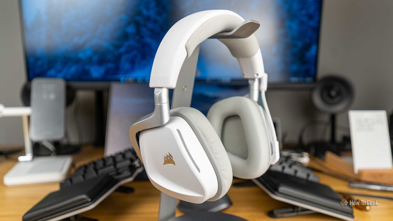 The CORSAIR Void Wireless V2 gaming headset in white sitting on a headphone stand on a desk with other gaming peripherals around it.