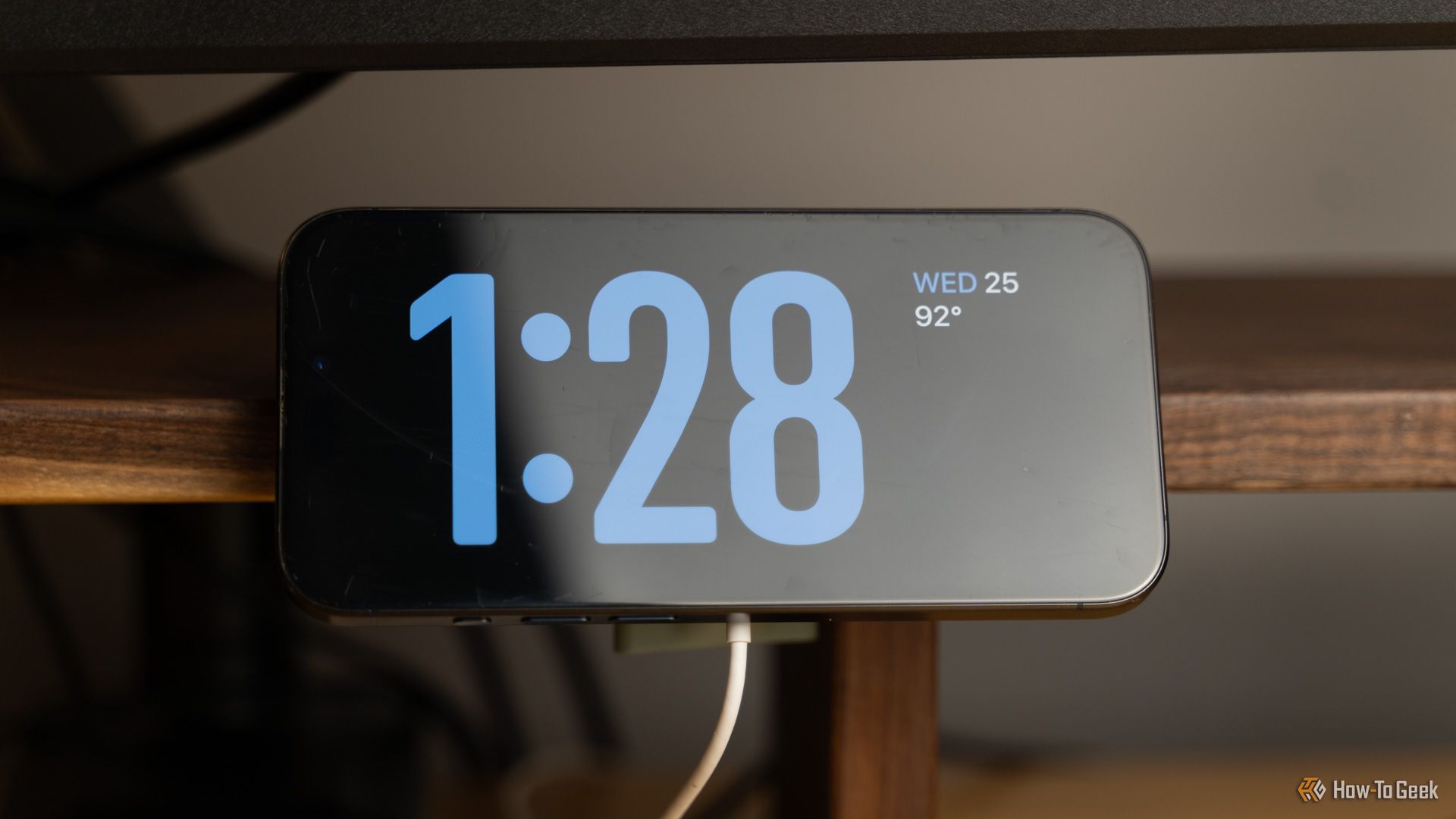 An iPhone mounted in Standby Mode on a 3D printed MagSafe charger mount that is clamped to a walnut desk shelf.