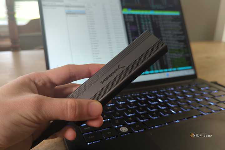 A person's hand holding a Sabrent external SSD drive in front of a Linux laptop.