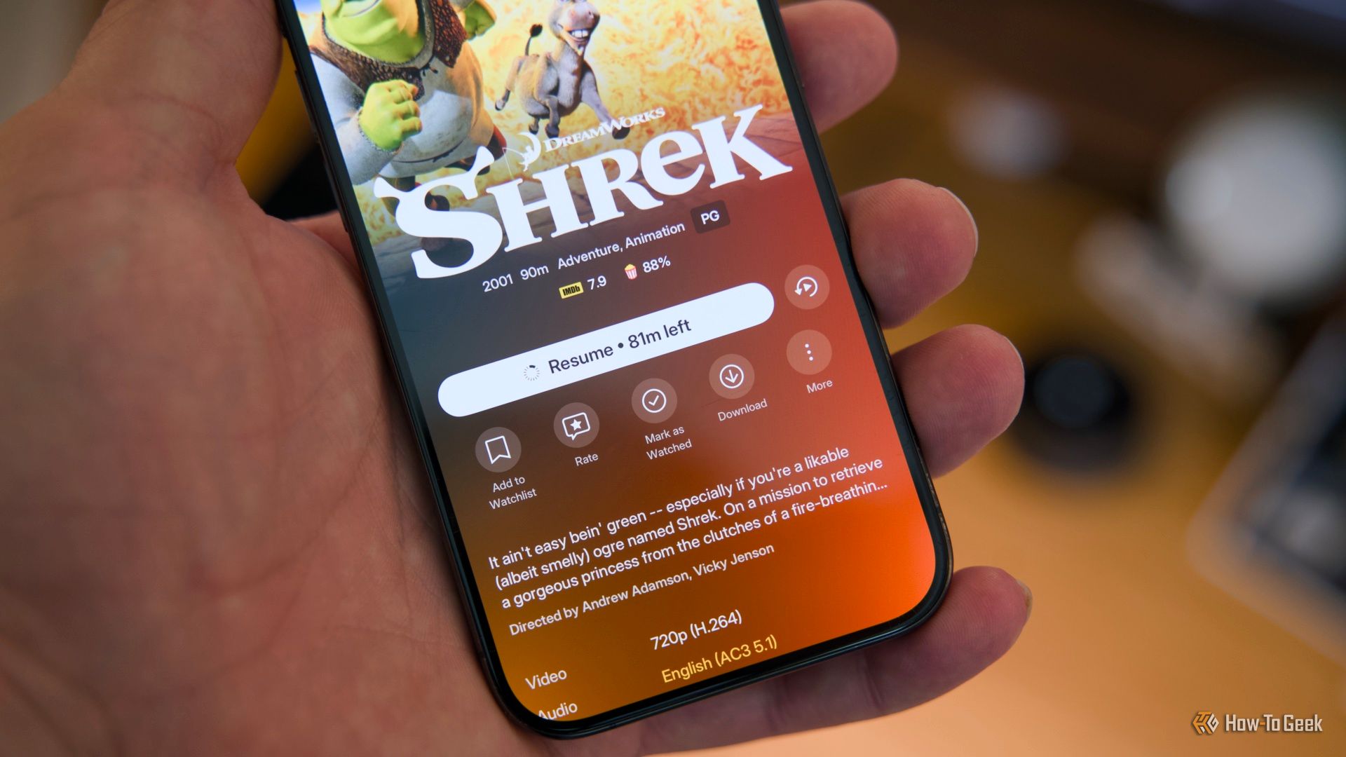 An iPhone 15 Pro held in a hand showing the Plex mobile app with the download icon visible on the Shrek movie.