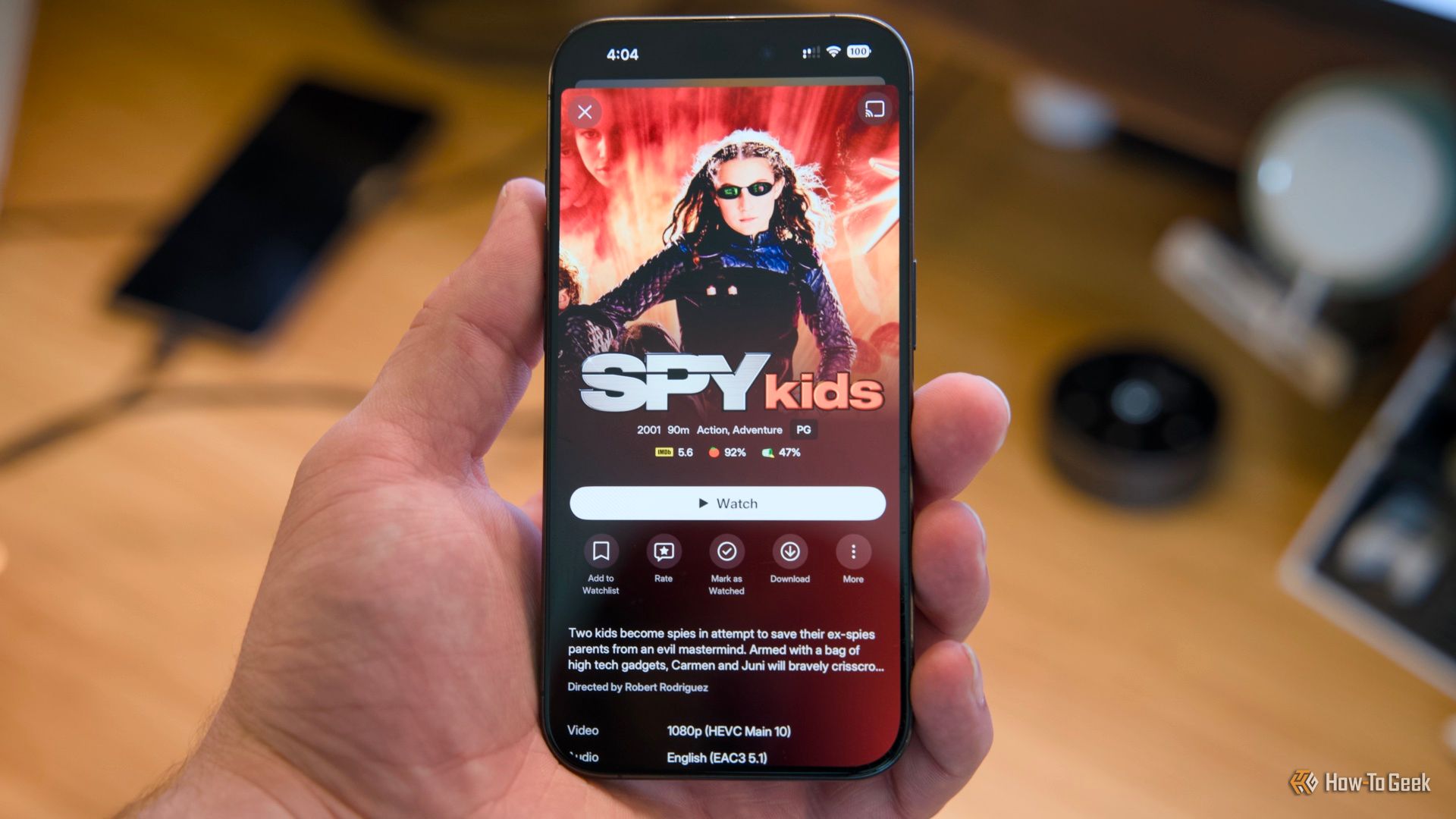 An iPhone 15 Pro held in a hand over a desk with the Spy Kids movie shown on the Plex mobile app.