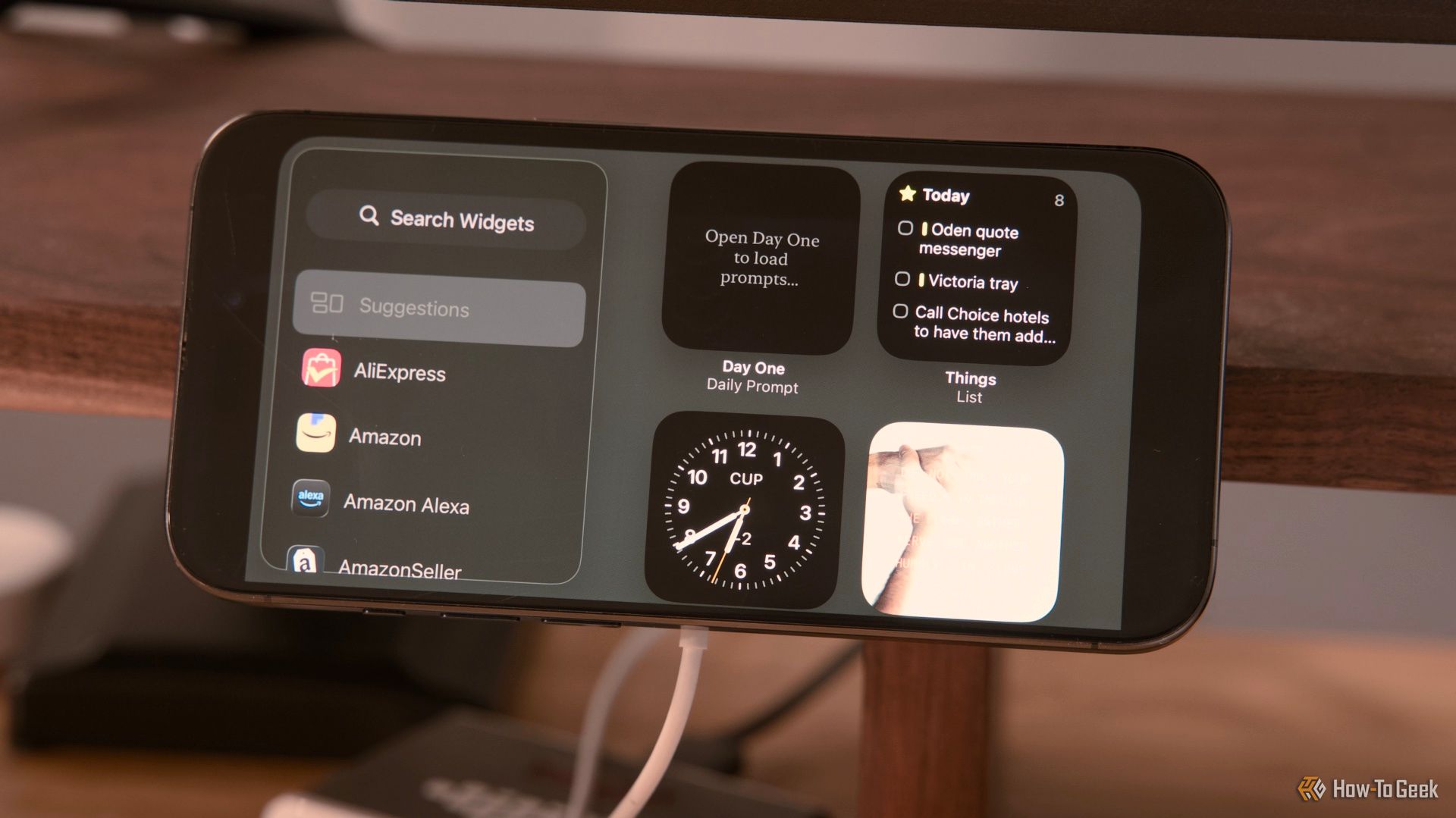 The widgets screen on an iPhone 15 Pro in StandBy mode mounted on a 3D printed MagSafe holder clamped to a desk.